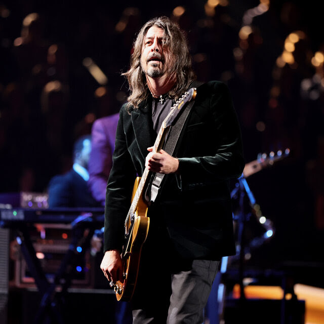 Dave Grohl, in a black jacket and gray pants, holds a guitar while standing onstage.