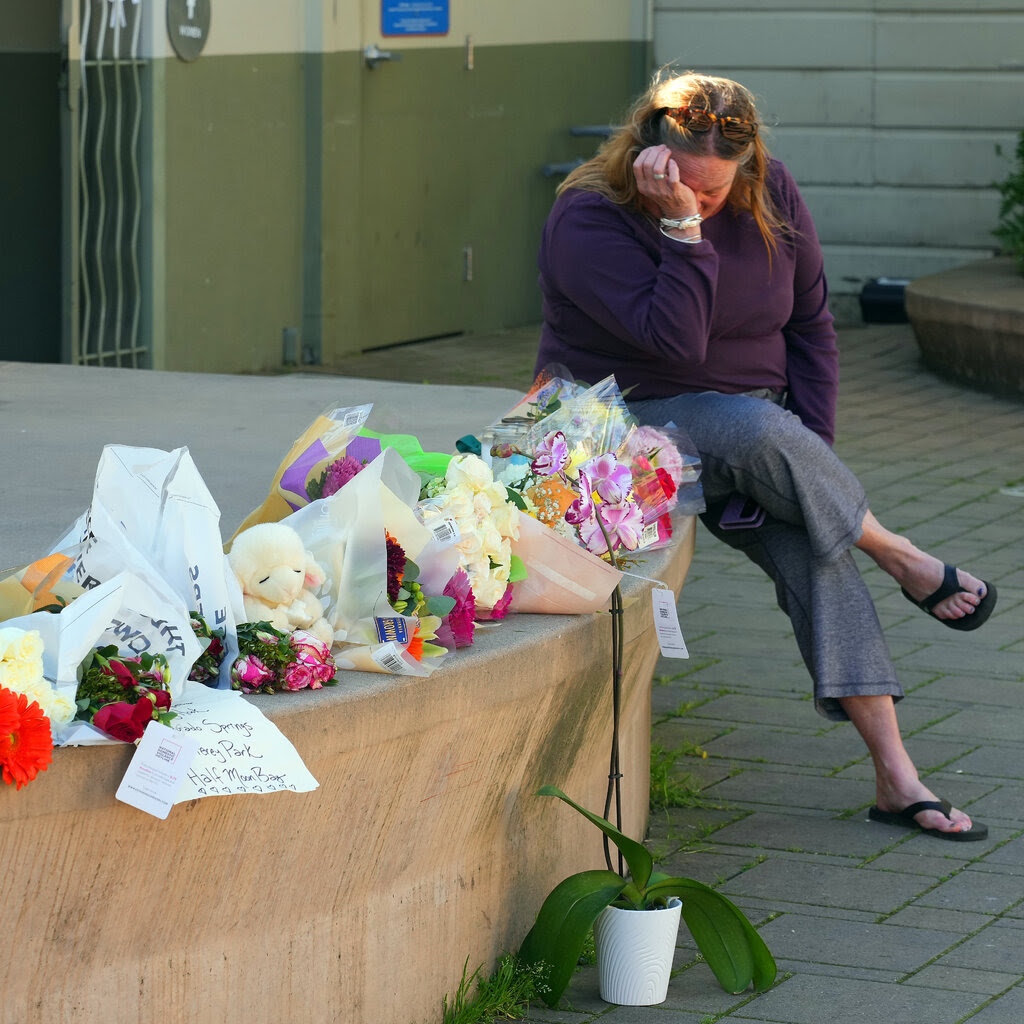 A woman pressing a hand against her face sitting on a ledge next to several bouquets of flowers.