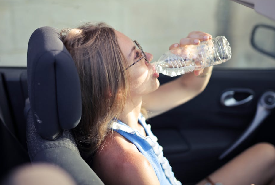 Free High angle side view of young woman in glasses and casual clothes drinking water from plastic transparent bottle while sitting in cabriolet with open roof in traffic jam in hot sunny day Stock Photo