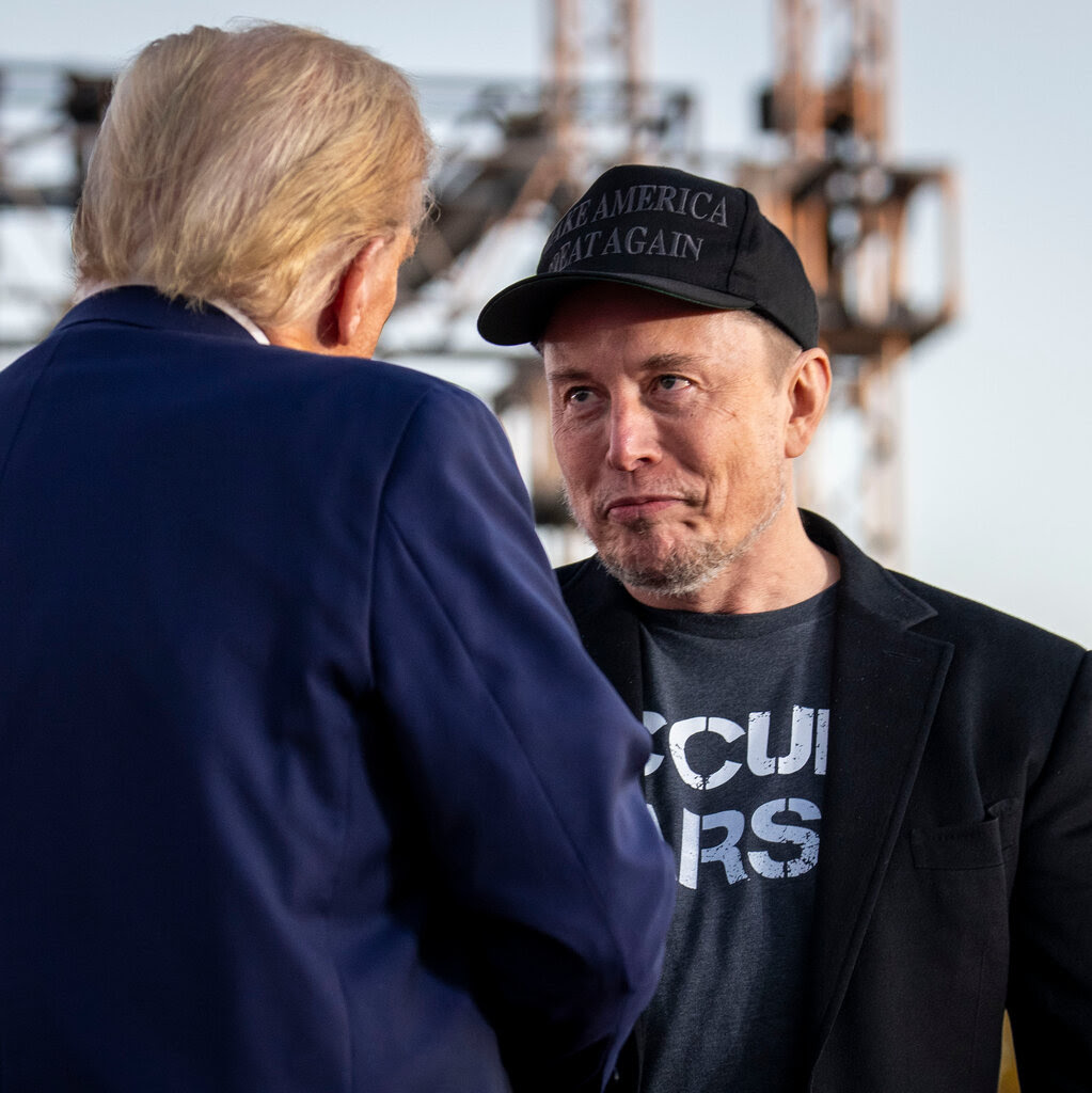 Donald Trump, seen from behind, with Elon Musk, who is dressed in black with a black MAGA baseball cap.