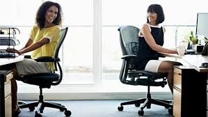 Two women in ergonomic office chairs at their desks, turning towards each other to chat. They are in a busy office environment, each with computers and papers on their desks, engaging in a friendly conversation while enjoying the comfort of their supportive chairs.