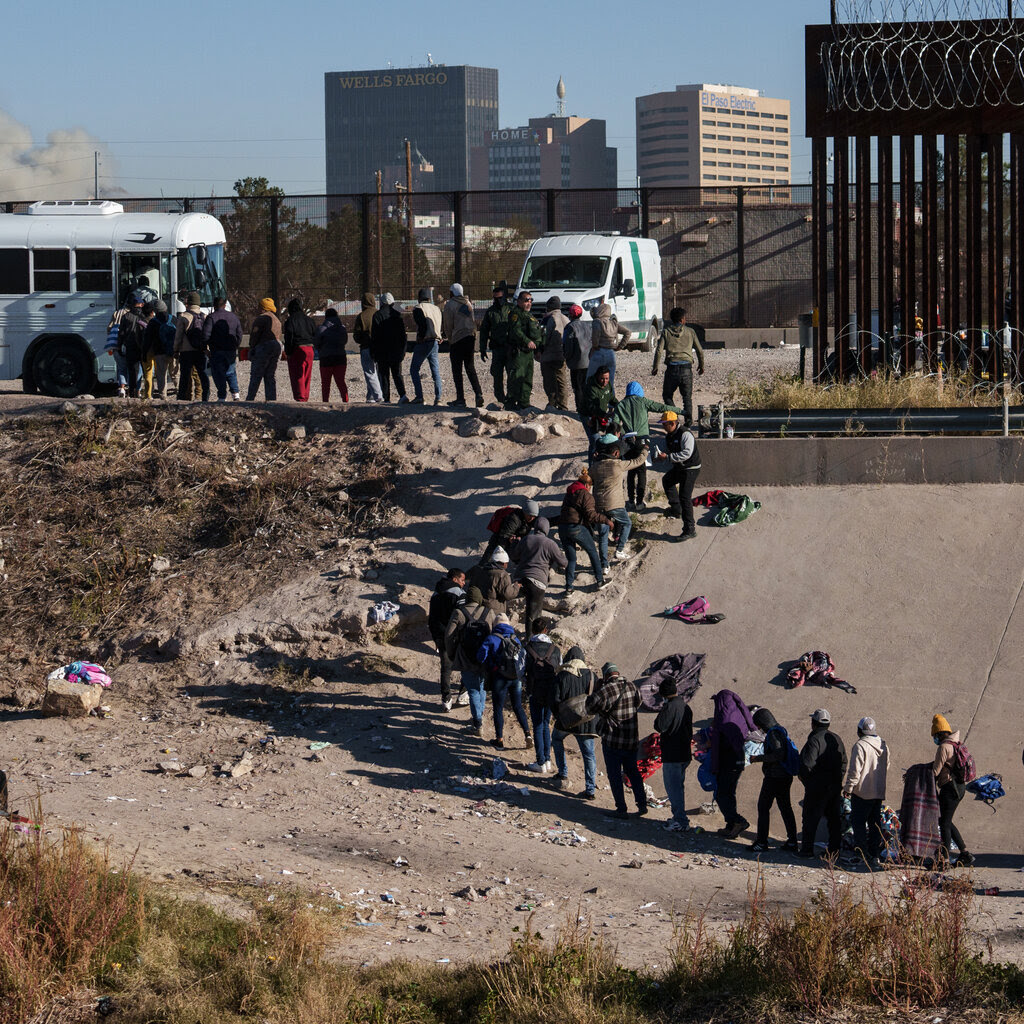 A long, serpentine line of people waiting to board a white passenger bus snakes its way down a steep concrete embankment and along a riverbank. 