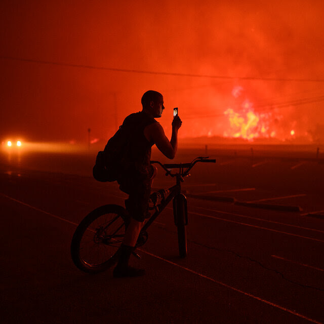 A man on a bike holds up a cellphone as the night sky behind him is turned orange by a large fire.