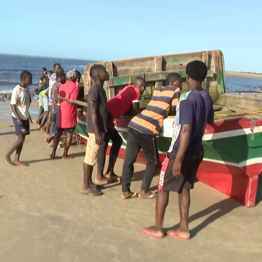 About a dozen people stand around a green-and-red wooden boat on a beach.