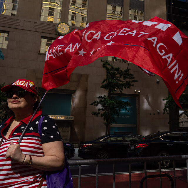 A woman in an American-flag shirt and a red MAGA hat holds a billowing red flag that reads Make American Great Again. 