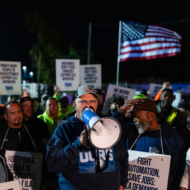 A man speaks into a megaphone surrounded by a crowd holding signs that say "Fight automation" and "ILA workers over machines."