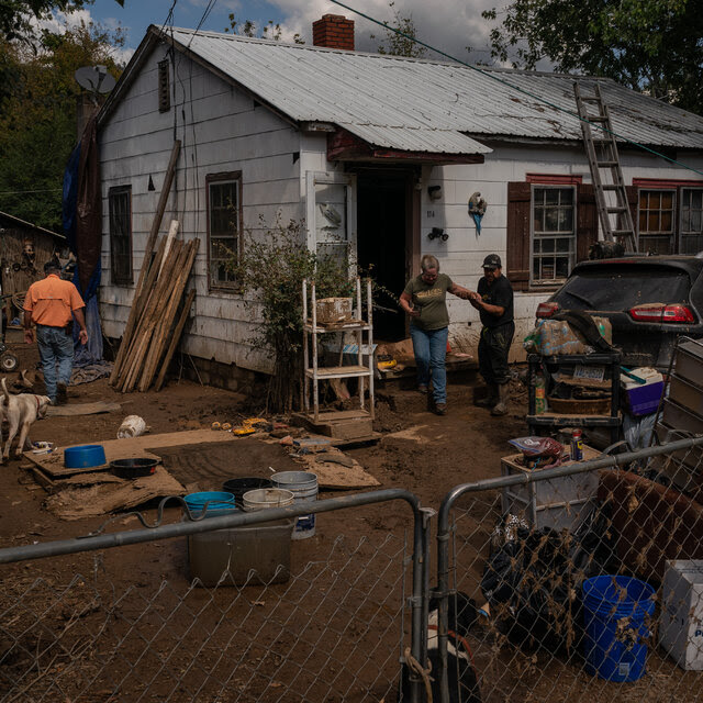 A man helps a woman to step from a damaged white house across a mud-caked yard filled with broken, muddy furniture and buckets.