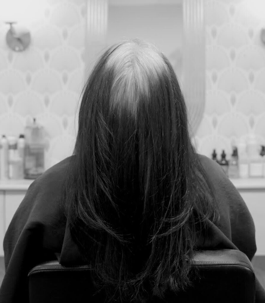 A woman sits in a salon in a black-and-white photo. Her hair is gray at the roots and dark and the ends.