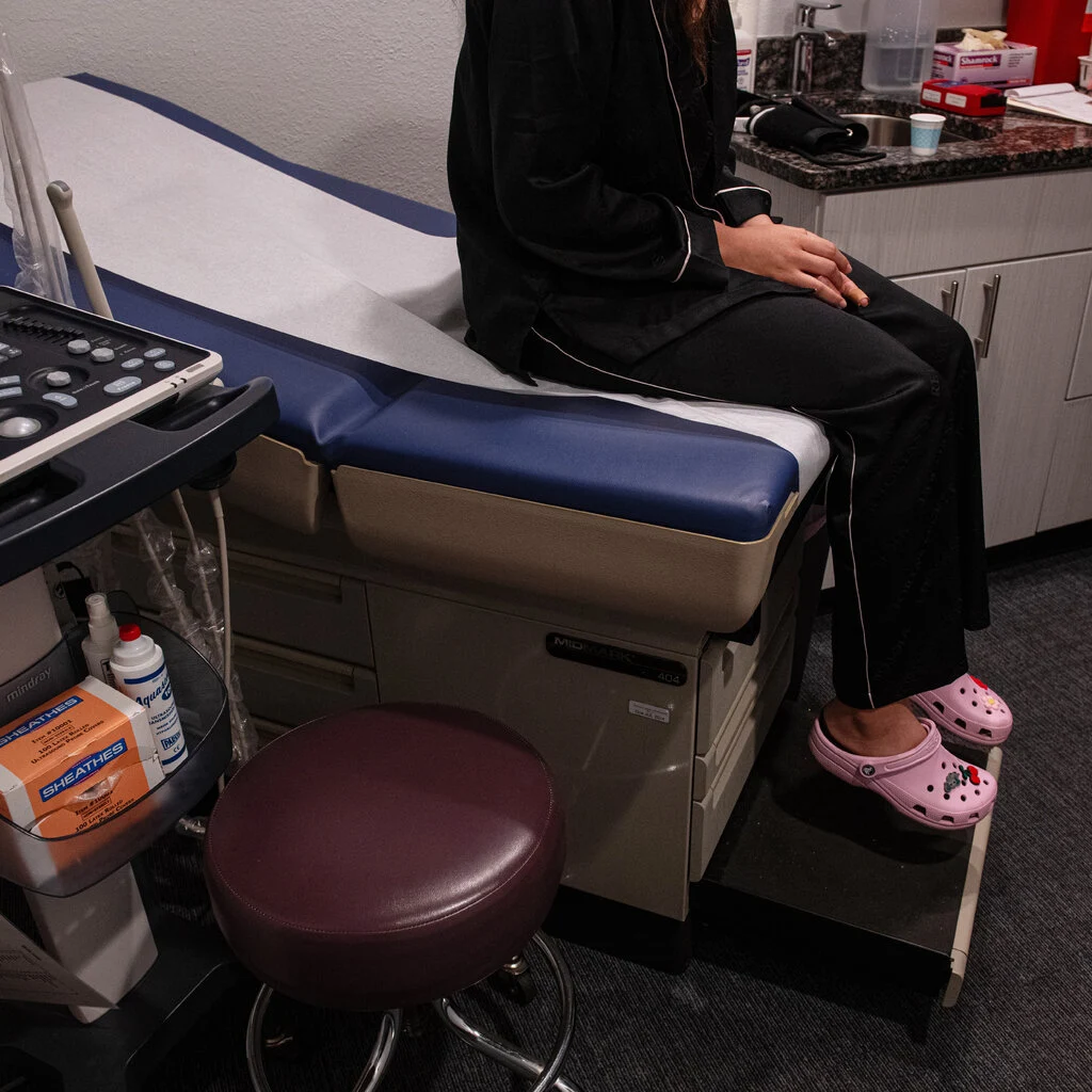 A patient sits on a medical bed while a clinic employee writes on a notepad next to her.