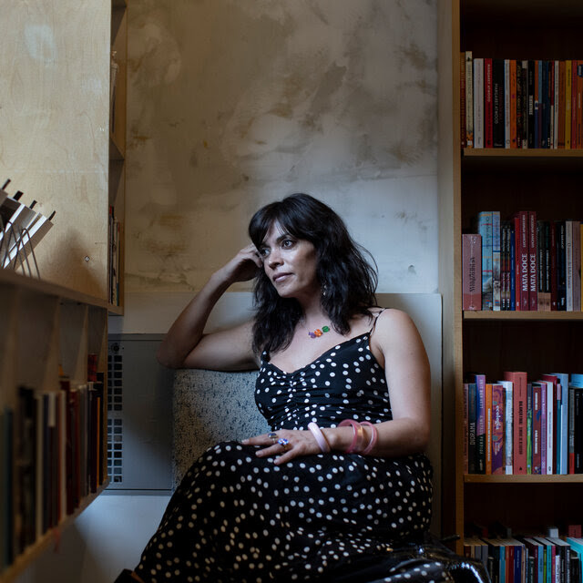 Argentine writer Camila Sosa Villada poses for a portrait at a bookstore in São Paulo. The room is in shadow, and she’s looking toward a window. 