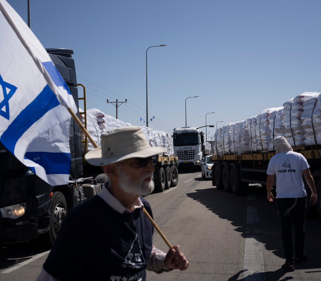 A man with an Israeli flag over his shoulder stands on a road covered with stopped trucks carrying aid intended for Gaza.