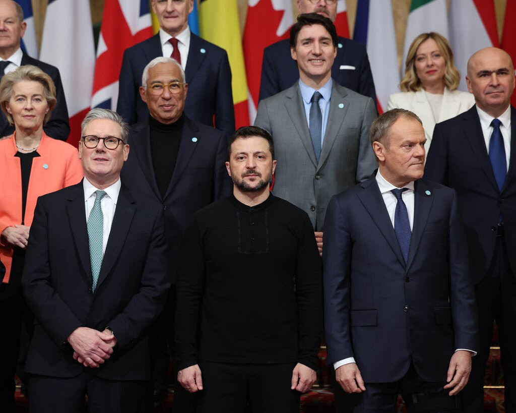 President Volodymyr Zelensky of Ukraine, wearing a black, collarless shirt, poses with European leaders in suits, standing in front of a row of flags.