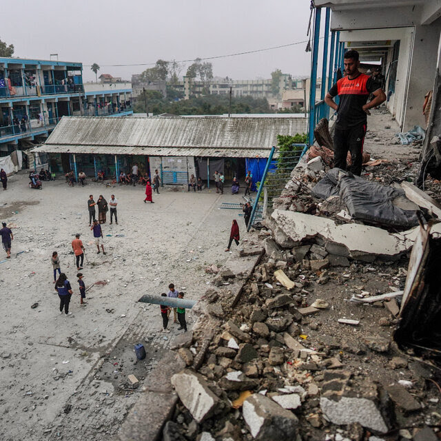 People standing in the courtyard of a damaged building. A man stands on an upper floor looking at rubble.