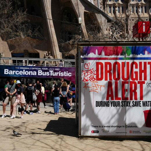 A couple uses a selfie stick to take a picture next to a banner warning tourists on drought alert in Catalonia, near Sagrada Familia basilica in Barcelona on April 13, 2024. From the Balearic Islands to the Canary Islands, Barcelona and Malaga, anti-mass tourism movements are multiplying in Spain. On the Canary Island Tenerife, activists started hunger strike on April 11 to demand a moratorium on mass tourism. (Photo by PAU BARRENA / AFP)