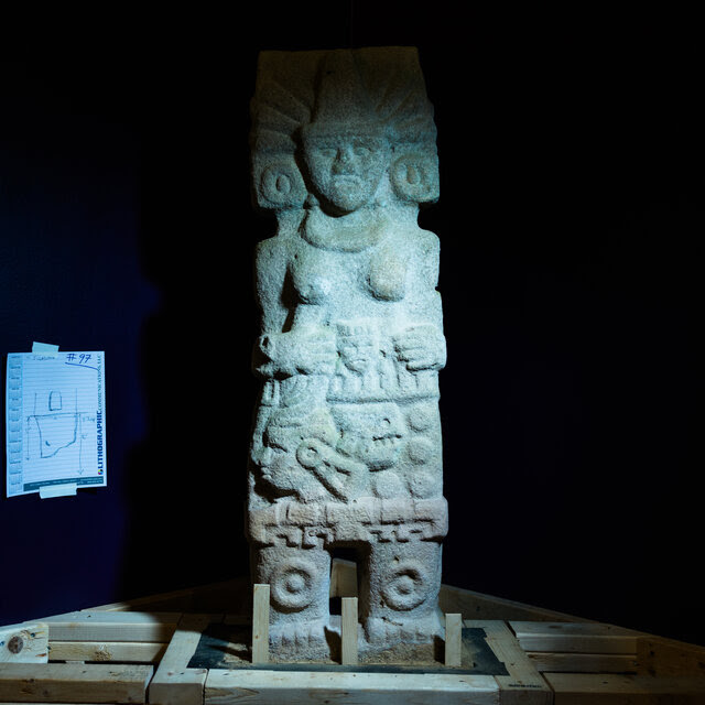 A six-foot statue of a woman with a large headdress and kneepads with a depiction of a severed head in her right hand, is placed on a wooden stand in a dark corner of a museum space.