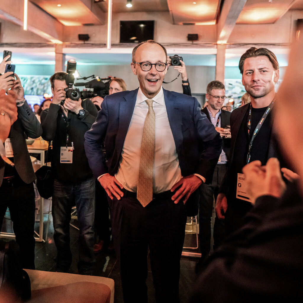 Friedrich Merz, wearing a suit, smiles while holding his hands on his hips as people take photos. 