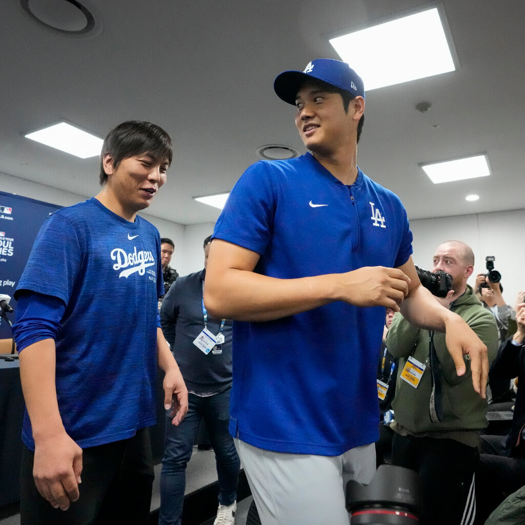 Two men in blue Los Angeles Dodgers shirts standing in a room full of photographers.
