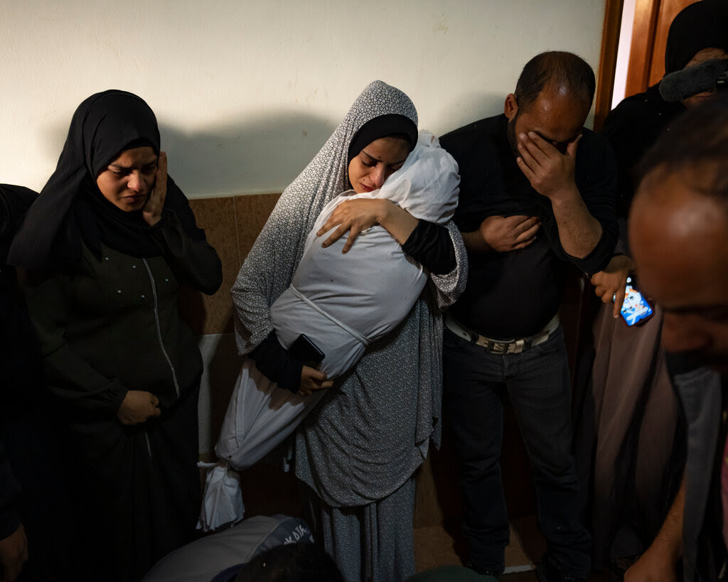 A woman clutches the shroud-wrapped body of a child as a man and a woman cry beside her.