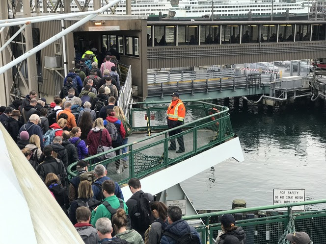 A large group of people leaving a ferry via a ramp at Bainbridge terminal, overseen by a crew member in an orange safety jacket