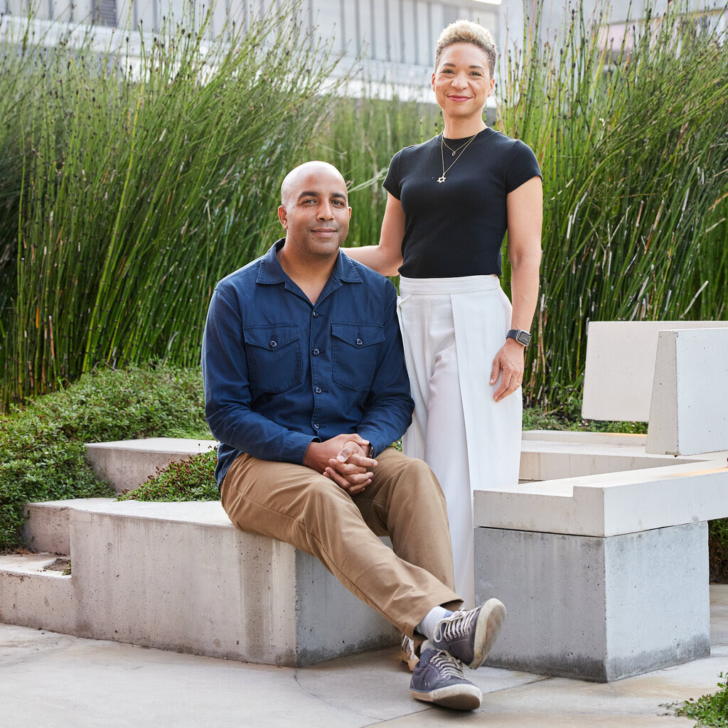 A man and woman pose for a photo. He is seated in a blue shirt and khakis, and she stands with her hand on his back, wearing white slacks and a black top.