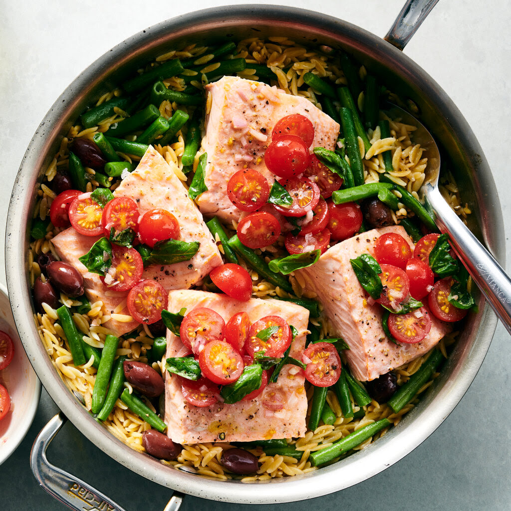 A skillet filled with orzo, green beans, salmon fillets and sliced cherry tomatoes is photographed from above. To the left is a small ceramic bowl filled with more sliced tomatoes.