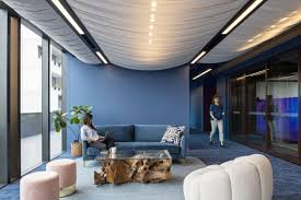  office lounge area with deep blue walls, stylish, comfortable seating arrangements including sofas and ottomans, and a distinctive wooden coffee table, all under a chic, wavy ceiling that adds a dynamic touch to the space.