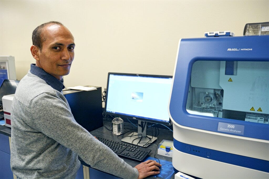Subas Malla, Ph.D. in lab at Uvalde center. He is wearing a gray sweater with a blue shirt underneath. He is standing in front of a computer monitor. 