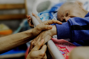 A frail, elderly man is reaching over a bed rail. Two caring hands are holding his.