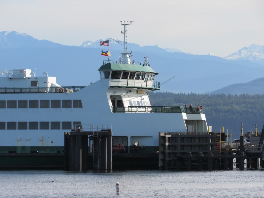 American and Pride flag flying atop a ferry docked at terminal with mountains in the background