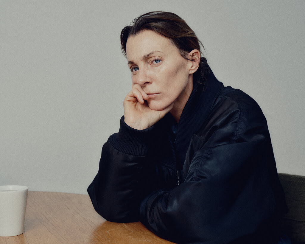 Phoebe Philo, in the blue bomber, sits at the table, her head resting in her hand. She appears deep in thought, 