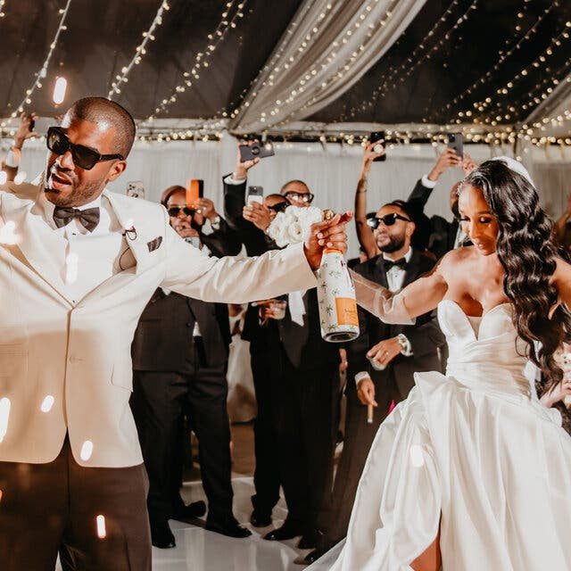 The bride and groom dance together on the dance floor. She is wearing a white sweetheart necklace dress with a slit front. He is wearing a white tuxedo jacket, black pants and sunglasses while holding a bottle of champagne. They are surrounded by standing guests snapping pictures, hanging string lights and a chandelier.