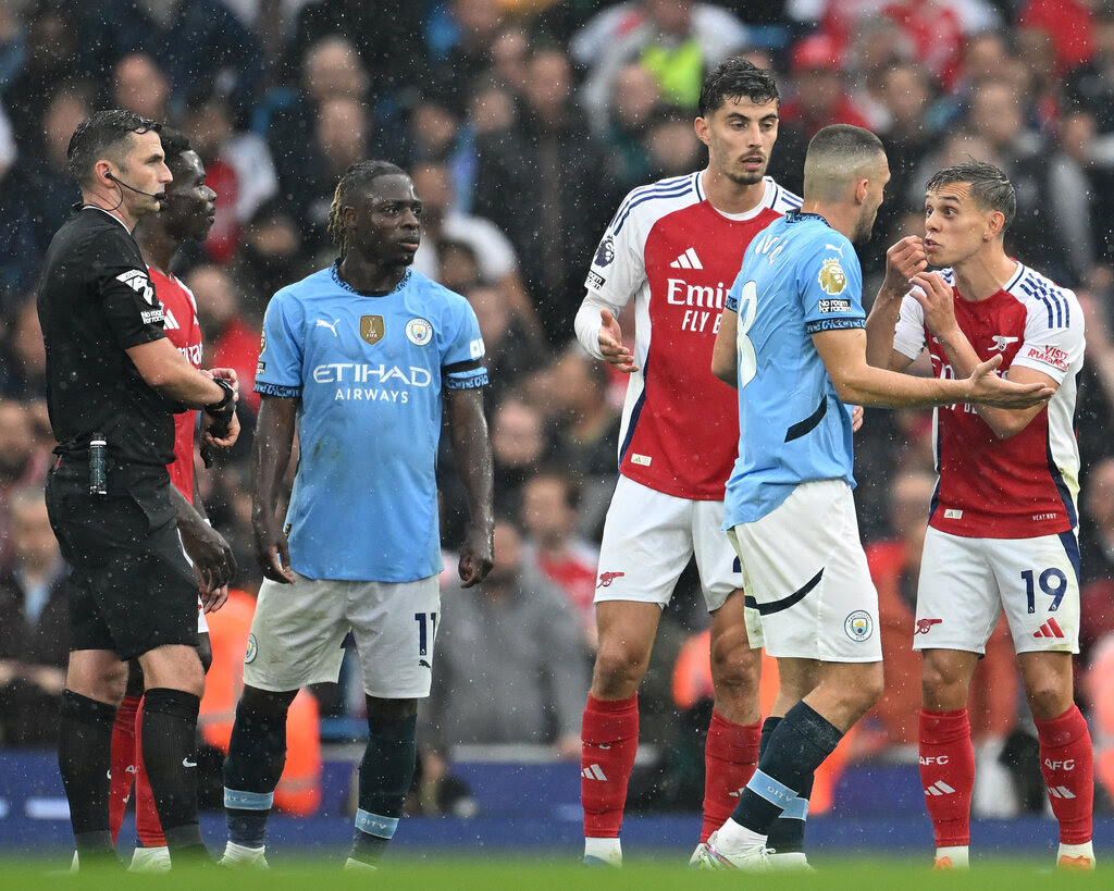 Multiple men are playing soccer in the rain, and they are fighting over a recent call by a referee who is also on the pitch. Out of focus behind the men is a crowd of fans and onlookers. 