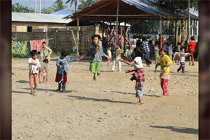 Children are jumping rope in the sunshine. There are other people in the background.