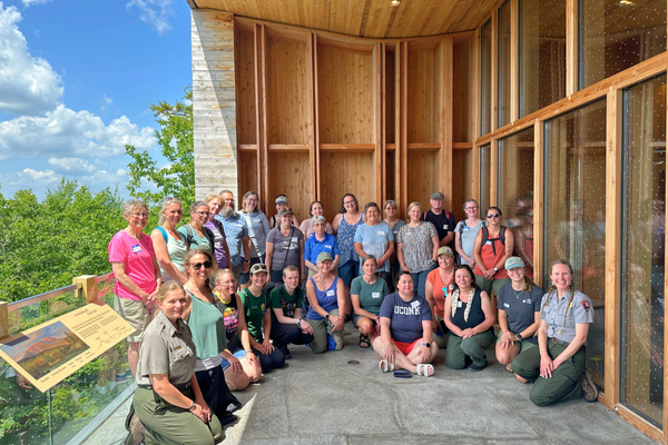 30 adults pose in a semicircle on the balcony of a modern wood building in the woods.