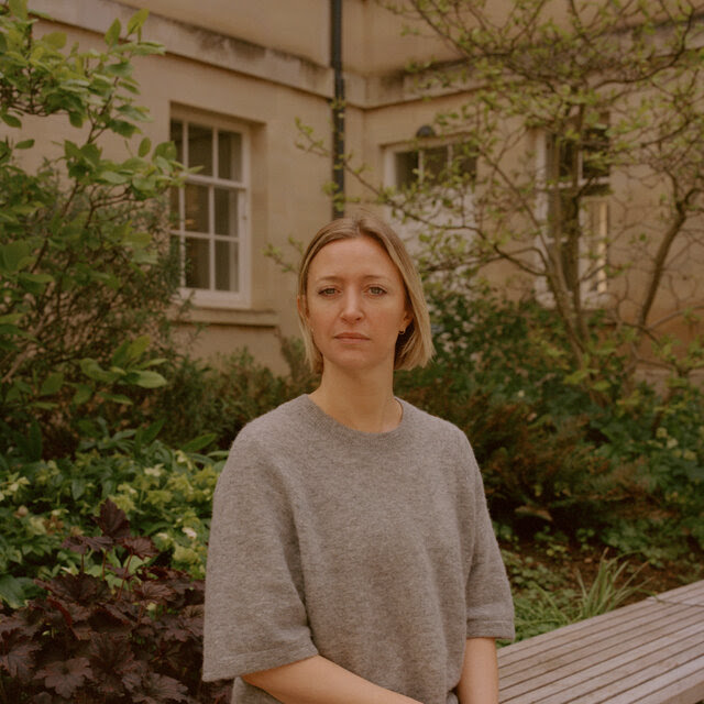 A portrait of Lucy Foulkes, who wears a gray sweater and black pants and sits on a bench in a garden area outside a building.