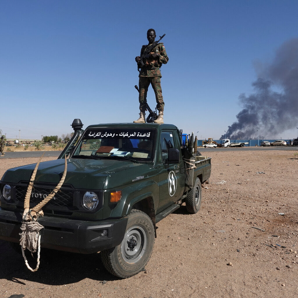 A man with his head covered stands on top of a pickup truck while holding a gun. In the distance, black smoke billows. 