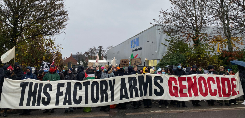  A large crowd of people bundled against the cold, some masked, hold a long banner: "THIS FACTORY ARMS GENOCIDE." A few small handmade signs are also visible: "Ceasefire now, end the occupation," "Jews against genocide," and a Palestinian flag. Behind them is the factory with a big "Eaton" logo. 