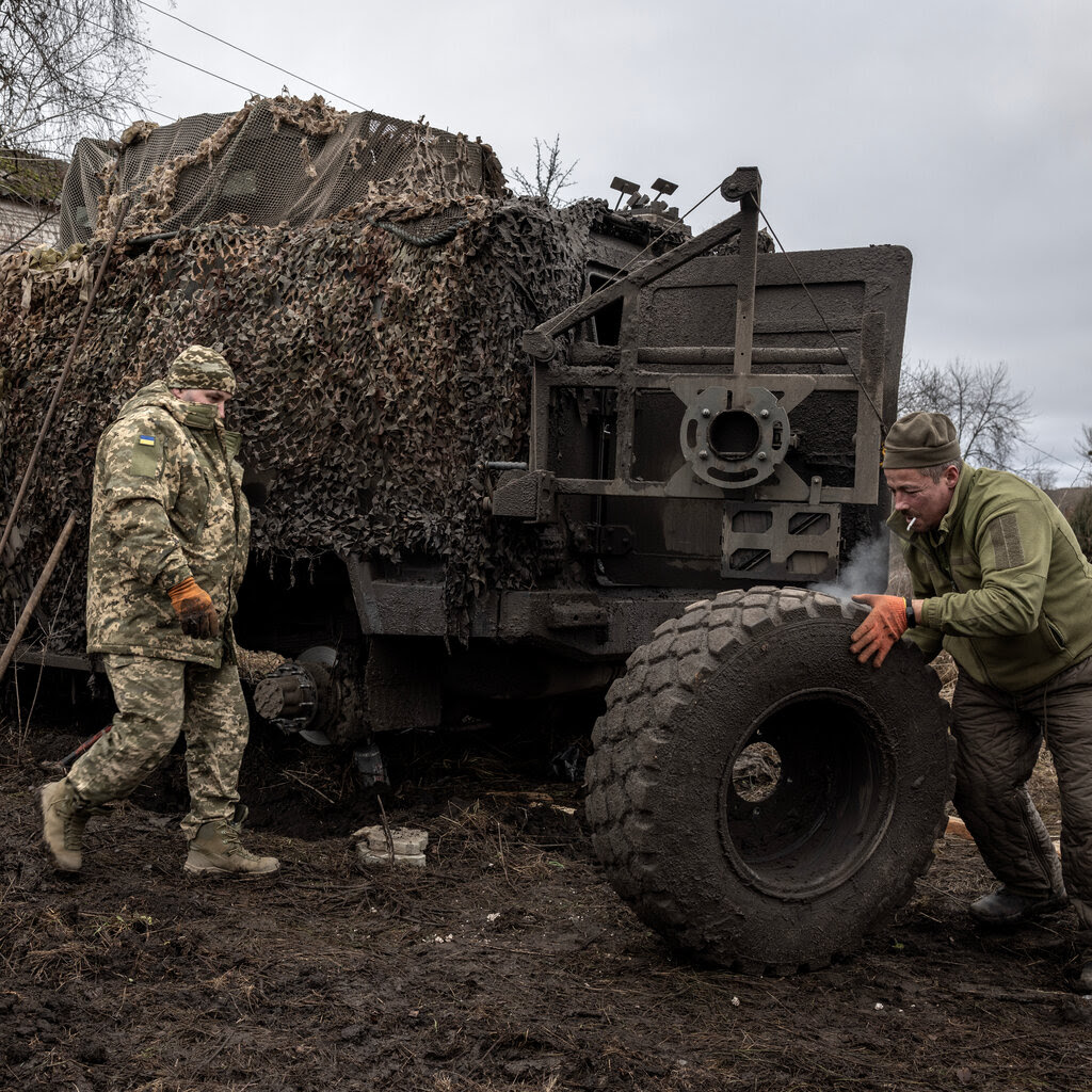 A man pushing a wheel toward a military vehicle as a soldier walks by. 