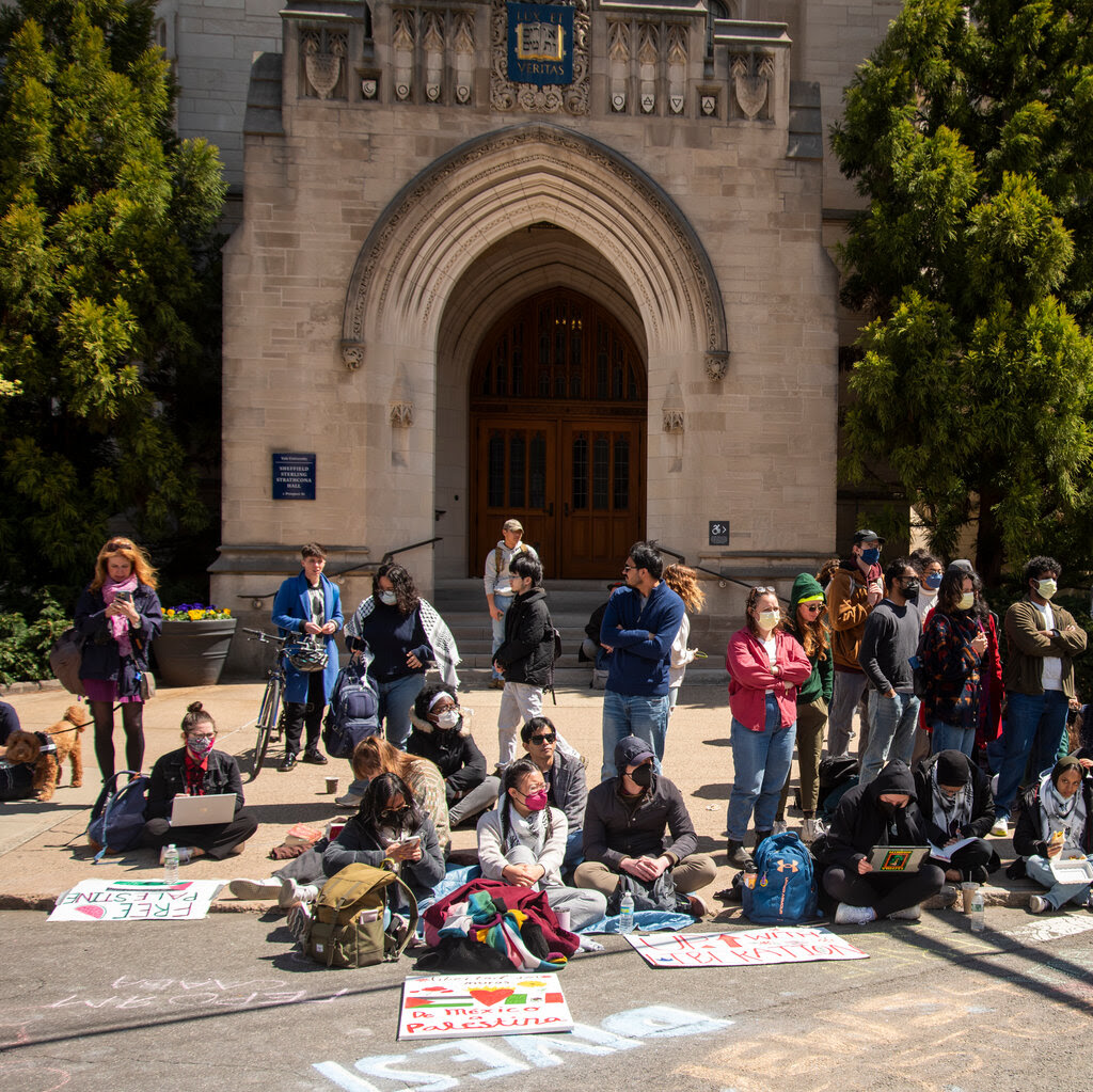 A group of students gathered in front of a university building. They are making signs. Many are wearing masks.