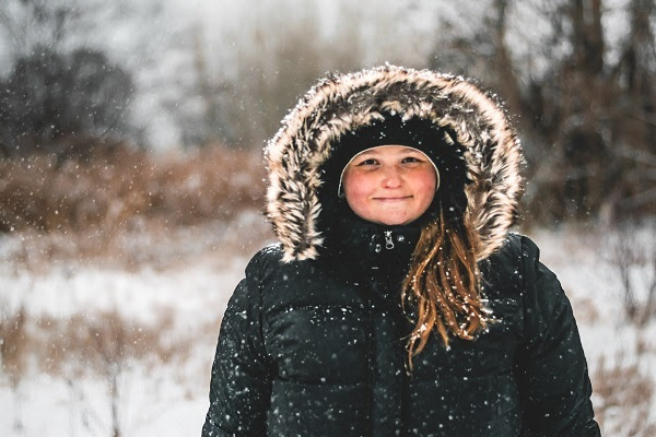 a young girl, brown hair spilling out from her hood, in a navy blue winter coat, smiles as light snow falls around her in a wooded area