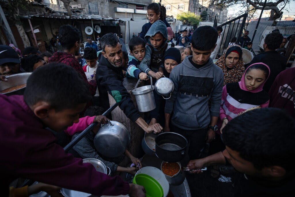 Children and some adults crowding to collect food while holding metal containers.