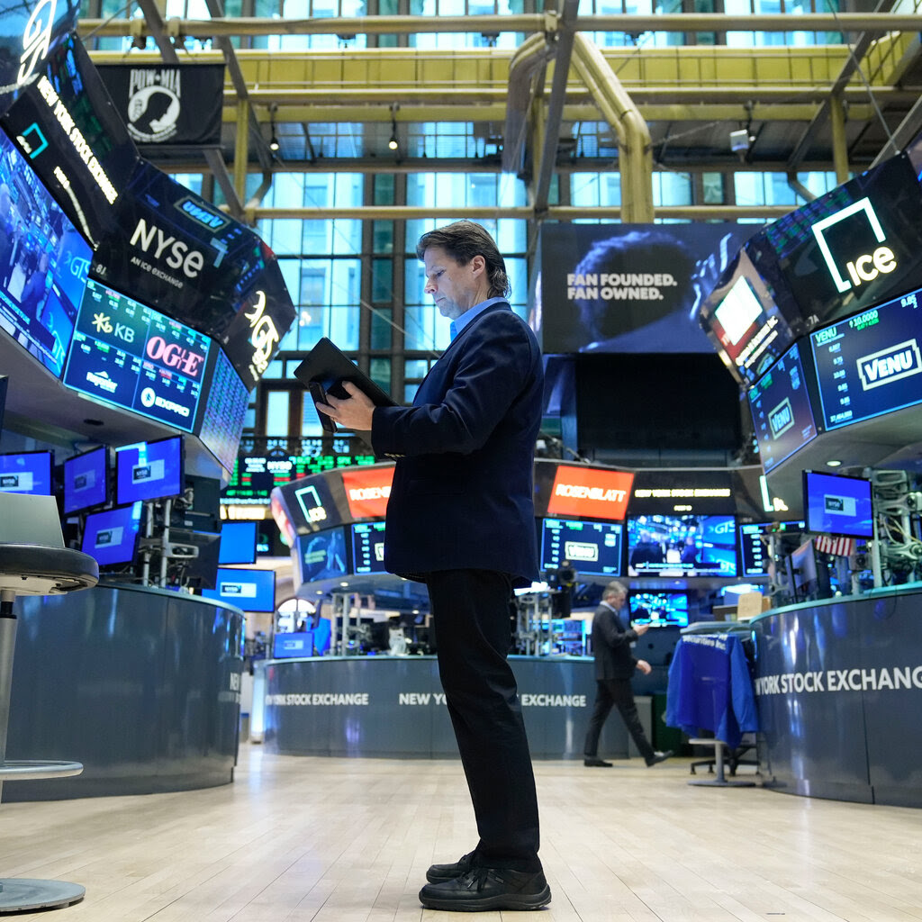 A man at the New York Stock Exchange holding a tablet standing amid large computer screens and TV monitors.