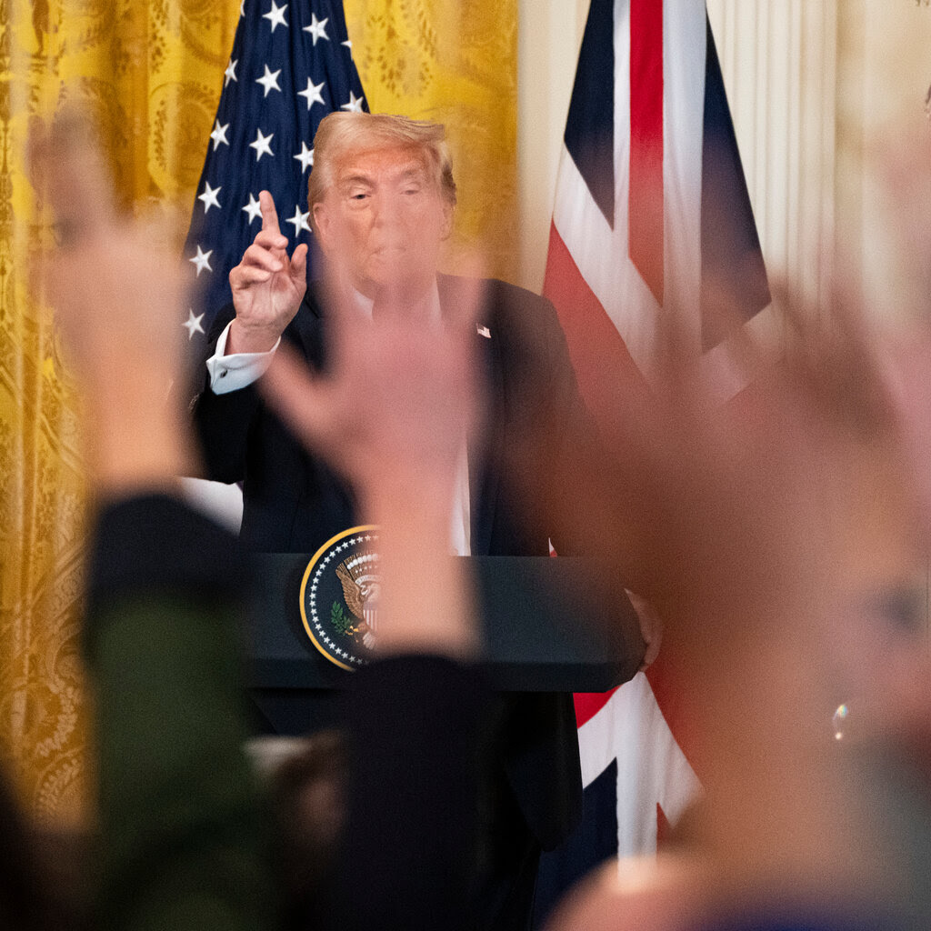 President Trump standing at a lectern and taking questions from reporters. Their hands are raised and appear out of focus.