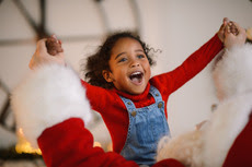 A young girl is smiling, her arms are opened wide into a champions pose by santa clause, back facing the camera
