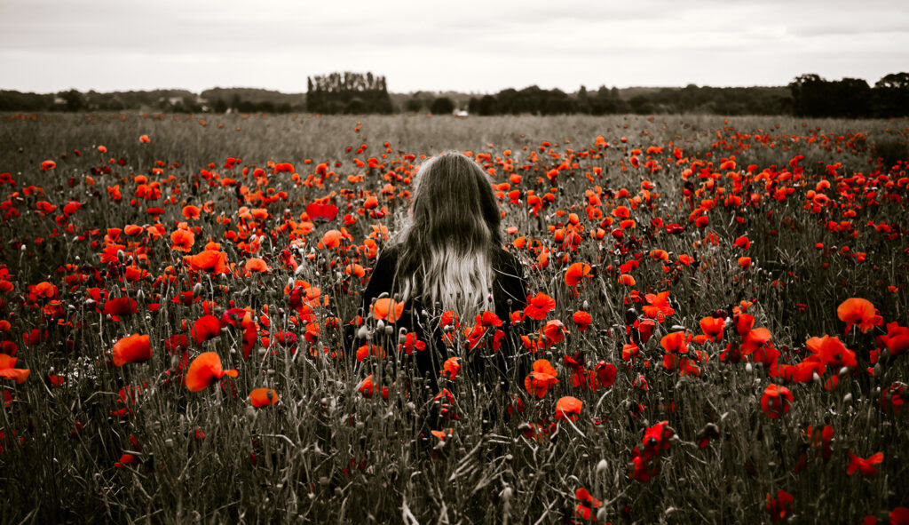 A woman with long black and grey-died hair, wearing black, is seen from behind standing in a field of tall orange flowers. The sky above is white and grey.