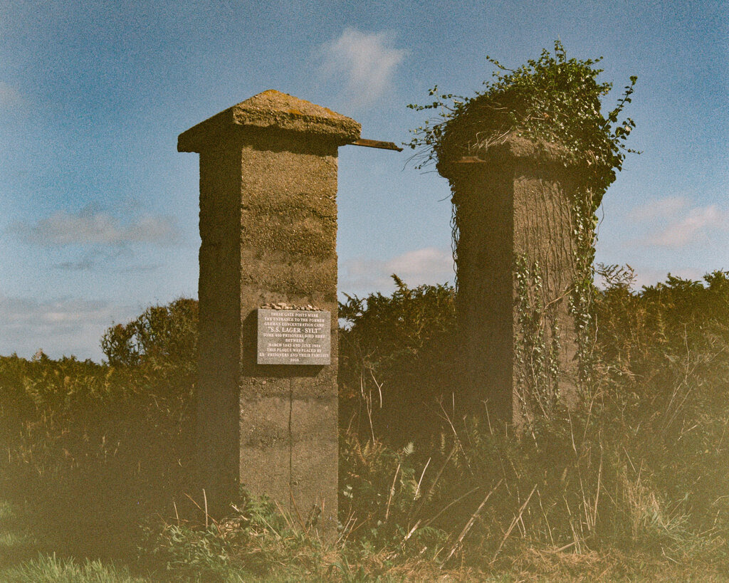 Two cement pillars stand in an overgrown field. There is a sign on one of them.