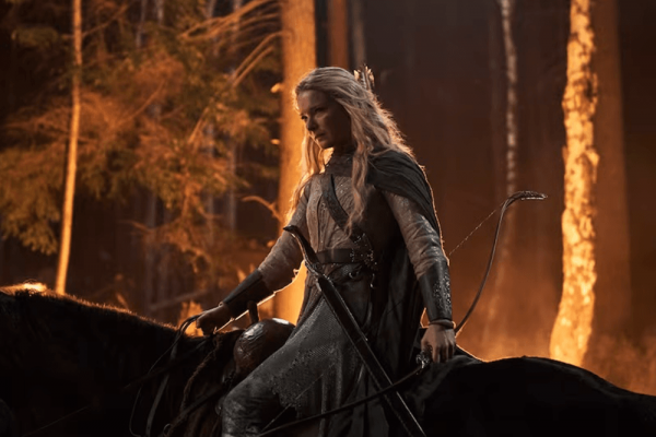 Woman in woodland armor holds bow and arrow and rides horse in forest.