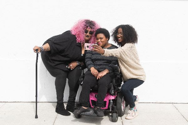 Three Black woman posing fo a selfie. Person on left has bright pink curly hair, wears dark glasses and holds a cane. Person in the middle has a short afro and sits in a wheelchair. The person on the right has dark shoulder length curly hair and is holding the phone out to take the photo