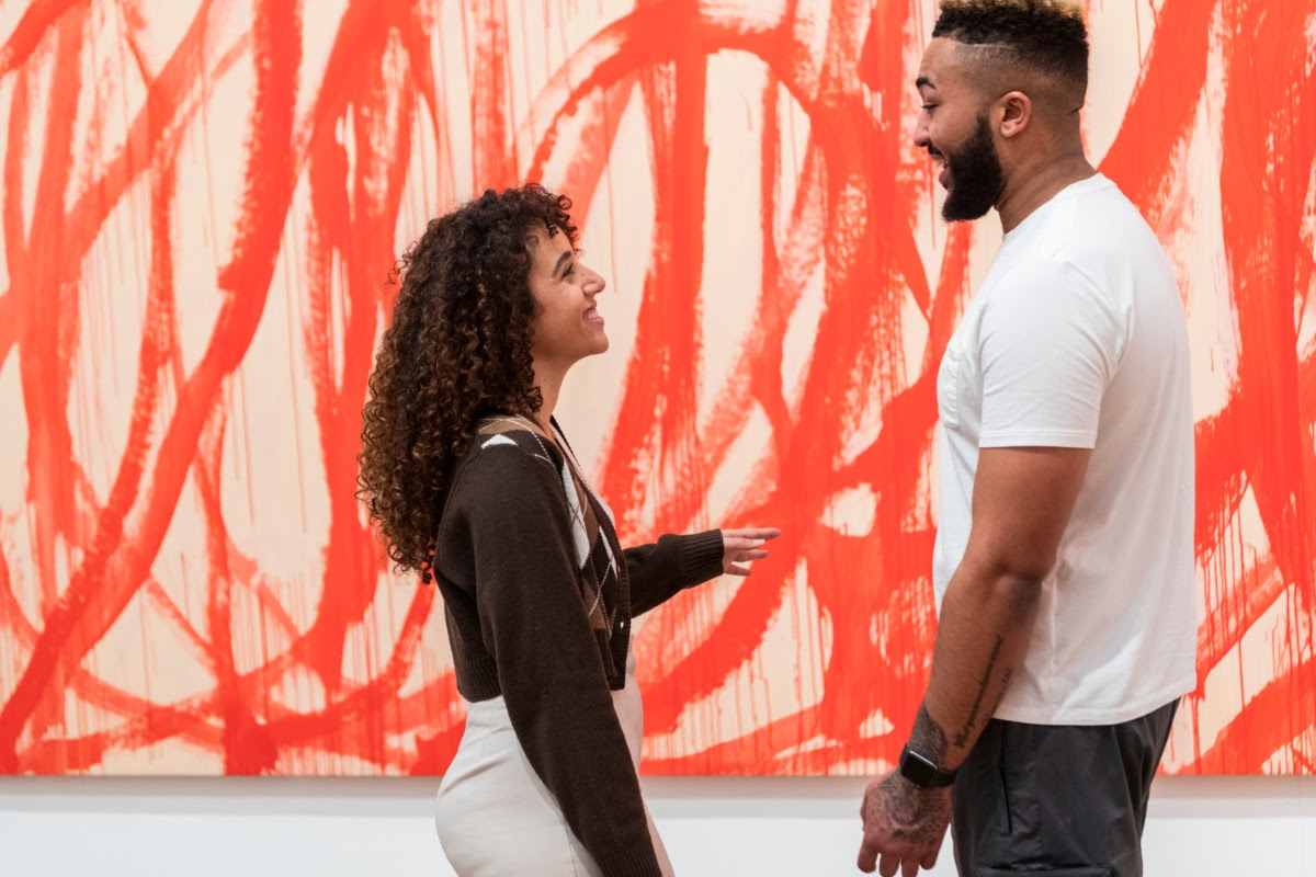 Just announced: Frida Kahlo, Tracey Emin and more! 9 Woman in brown top looks at man in white tshirt in front of a large scale abstract canvas with orange paint