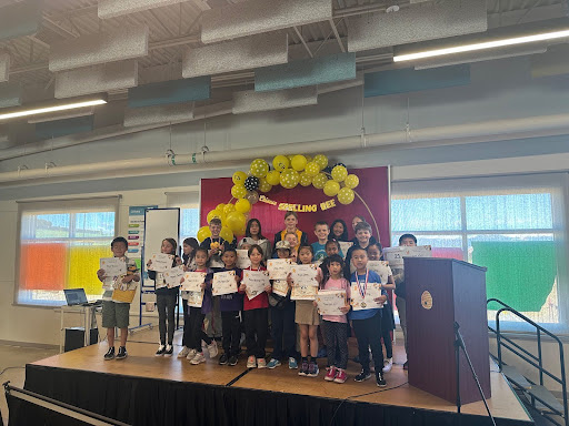 A group of children stand on a stage holding certificates, with a "Spelling Bee" banner and balloon decorations behind them. They are inside a brightly lit room with large windows.